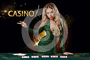 Lady in green dress is showing inscription casino, leaning on playing table with cards on it, posing on black background