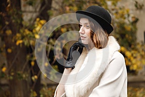 A lady in a hat, gloves and a light coat is looking out, holding her hand by the face. Retro. Outdoors.