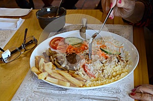 A lady having lunch at the restaurant with rice, farofa, beef steak with onions, grilled on a plate, french fries and a tomato,
