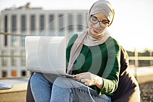 Lady in hijab putting charge cable into laptop outdoors