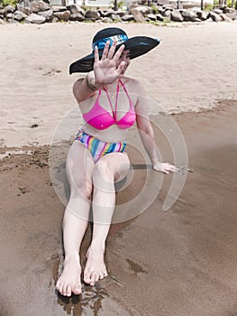 A lady in a large sun hat and bikini declines to have her photo taken at the beach, preferring to go incognito and not have