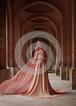 Lady in long pink dress in road coat with a hood is walking along old castle