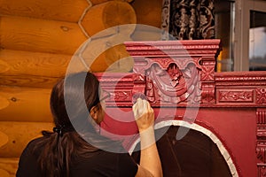 Lady painting splendid carved ornaments on cupboard made of wood in red with brush looking at upper part in workshop for