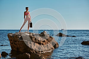 Lady in pink bikini with suitcase standing on rock. Beach vacation. Summer holiday, Time for travel. Adventure trip.