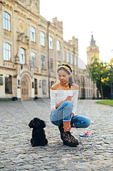 Lady playing with a cute black dog breed poodle toy on the cobblestones in the park on a background of beautiful landscape