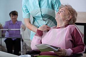 Lady reading a newspaper in common room of professional nursing home with helpful social worker supporting her