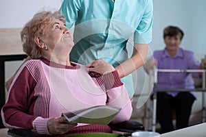 Lady reading a newspaper in common room of professional nursing home with helpful social worker supporting her