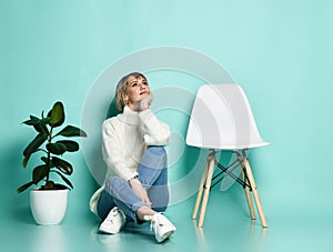 Lady in sweater, jeans, sneakers. Smiling, propping her face, sitting on floor between chair and ficus, posing on blue background