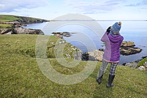 A lady in winter clothes and bobble hat looks with binoculars at a coastline landscape of rocks and sea.Shetland.