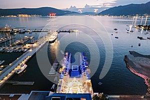 Laid tables on the pier near a festively lit stage in front of a long seaside pool. Drone