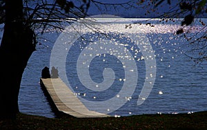 Lake with 2 vacationers on the bathing jetty in front of the low-lying sun