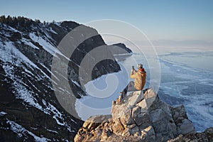 Lake Baikal at winter. Man sitting on a cliff and taking photo on smartphone. Frozen Baikal lake. Deepest and largest fresh water