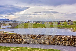 Lake with bank of stone and arched bridge under sky filled with clouds