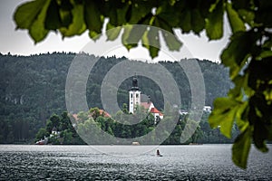 Bled lake with a Church of the Assumption of Mary view, and a girl on a paddleboard, in a frame of leaves in summer day time.