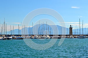 Lake landscape with sailboats, lighthouse, blue water, white clouds, mountains in haze. Lake Garda in Italy.