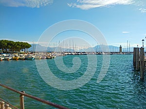 Lake landscape with sailboats, lighthouse, blue water, white clouds, mountains in haze. Lake Garda in Italy. Vacation