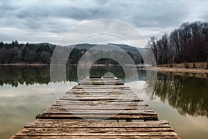 Jezerní molo na večer s kopci na pozadí. Odraz lesa v zelené vodě s modrou zataženou oblohou. Rybník u fores