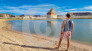 Lake Powell - Rear view of man walking at lakeshore looking at solitary rock formation Lone Rock in Wahweap Bay, USA