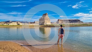 Lake Powell - Rear view of man walking at lakeshore looking at solitary rock formation Lone Rock in Wahweap Bay, USA