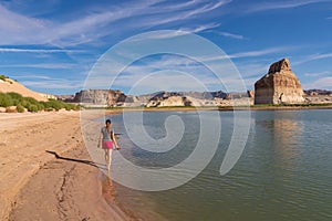 Lake Powell - Rear view of woman walking at lakeshore looking at solitary rock formation Lone Rock in Wahweap Bay, USA