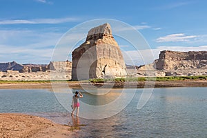 Lake Powell - Rear view of woman walking at lakeshore looking at solitary rock formation Lone Rock in Wahweap Bay, USA