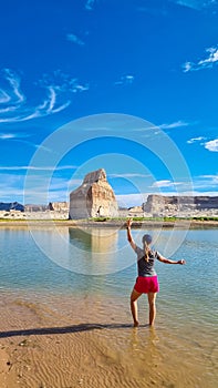 Lake Powell - Rear view of woman walking at lakeshore looking at solitary rock formation Lone Rock in Wahweap Bay, USA