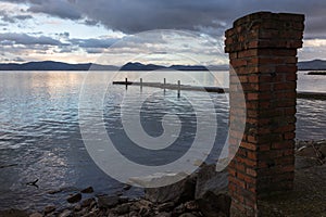 A lake shore at dusk, with an old brick column in the foreground