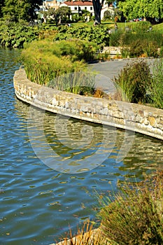 Lakeside cement wall with twisting and winding wall and sidewalk with shrubs and plants on outskirts of pond and water