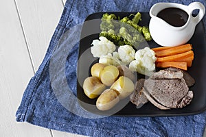 Lamb roast Sunday lunch dinner with vegetables on a aquare black plate, with gravy jug, on a blue tea towel.