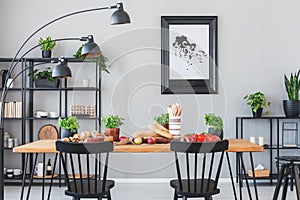 Lamp above black chairs and wooden table with food in grey dining room interior with poster