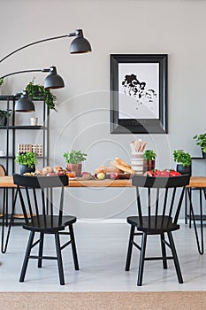 Lamp above black chairs and wooden table with food in grey dining room interior with poster. Real photo
