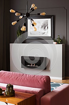 Lamp above pink settee in grey modern living room interior with poster above fireplace