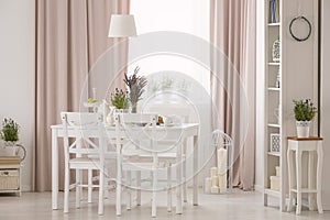 Lamp above table and white chairs in pink dining room interior with plants and drapes. Real photo