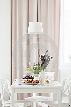 Lamp above white table and chairs in pastel dining room interior with plants and drapes. Real photo