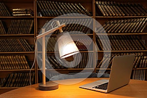Lamp and laptop on wooden table near shelves with collection of vintage books in library