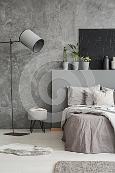 Lamp and stool next to bed with pillows and bedhead in grey bedroom interior with poster. Real photo photo