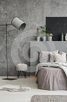 Lamp and stool next to bed with pillows and bedhead in grey bedroom interior with poster. Real photo