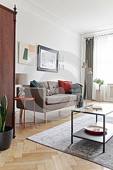 Lamp on table next to sofa in bright living room interior with posters and grey carpet. Real photo