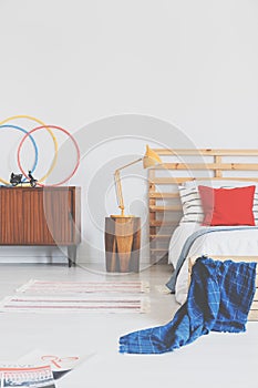 Lamp on wooden stool next to bed with red cushion and blue blanket in bedroom interior. Real photo