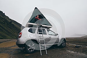 Landmannalaugar, Iceland - August 2018: Young woman sitting on a ladder next to offroad car with tent on the roof