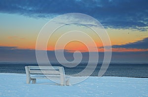 Landscape with bench on snowy shore at sunset.