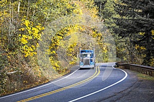 Landscape with blue big rig semi truck and bulk semi trailer running on the winding road with autumn forest on the sides