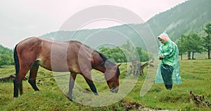 Landscape cloudy scenic view. rain in background. Woman walks on meadow in raincoat looks at brown horse.Background are