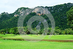 Landscape of a golf field with green grass, water hazard, trees, and mountain under cloudy blue sky 2