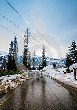 A landscape of a hilly highway after rain with snow capped houses by the side . Snow capped mountain and pine trees in the