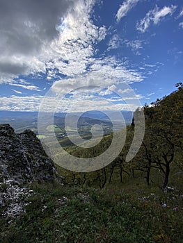 Landscape of a mountainous area with green slopes, trees, spring flowers and a blue sky with white clouds.