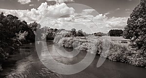 Landscape with a river next to a field and a farm with clouds in the background, black and white