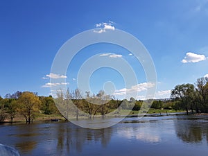 Landscape. The river on which are displayed green trees that grow in the shoreline. Blue sky with shelves. The soil is covered wi