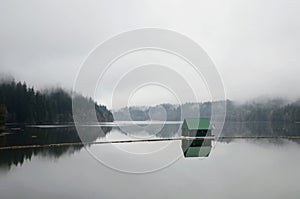 Landscape shot of a lake with a small green floating house in the middle during a foggy weather
