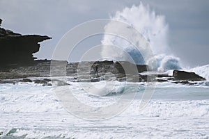 Landscape shot of a surfer standing on the rocky shore of the sea with waves crashing the rocks